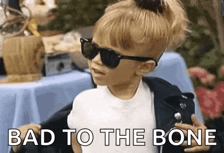 a little girl wearing sunglasses and a leather jacket is standing in front of a table and says `` bad to the bone '' .