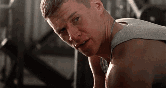 a man wearing a gray tank top and a necklace looks at the camera