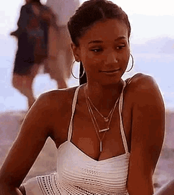 a woman is sitting on the beach wearing a white tank top and a necklace .