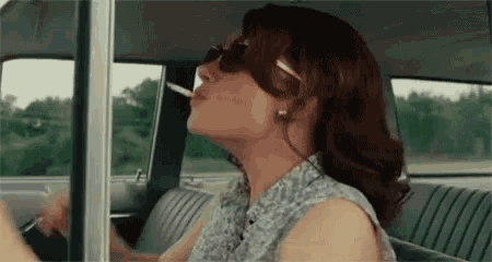 a woman is smoking a cigarette in a car while wearing sunglasses .