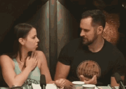 a man and a woman are sitting at a table . the woman is pointing at the man 's arm .