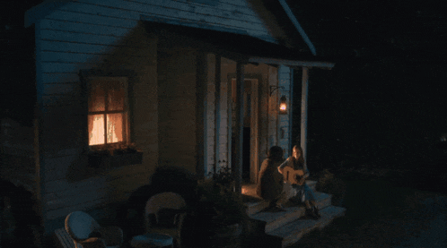 a woman sits on the porch of a house at night playing a guitar