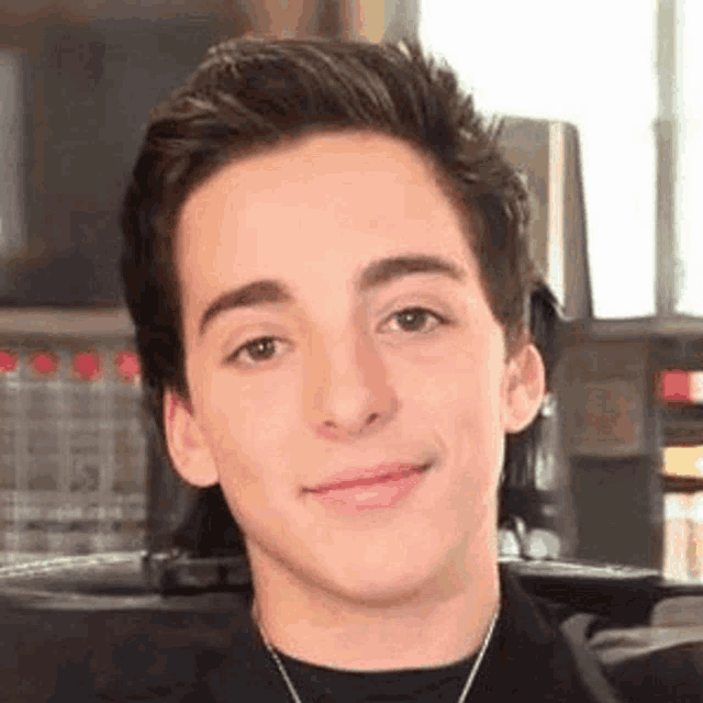a young man wearing a black shirt and a necklace is looking at the camera .