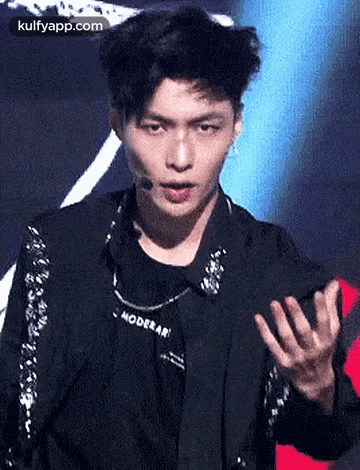 a young man wearing a black jacket and a necklace is standing on a stage with his hands outstretched .