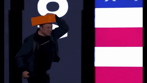 a man wearing a brick on his head stands in front of an american flag .