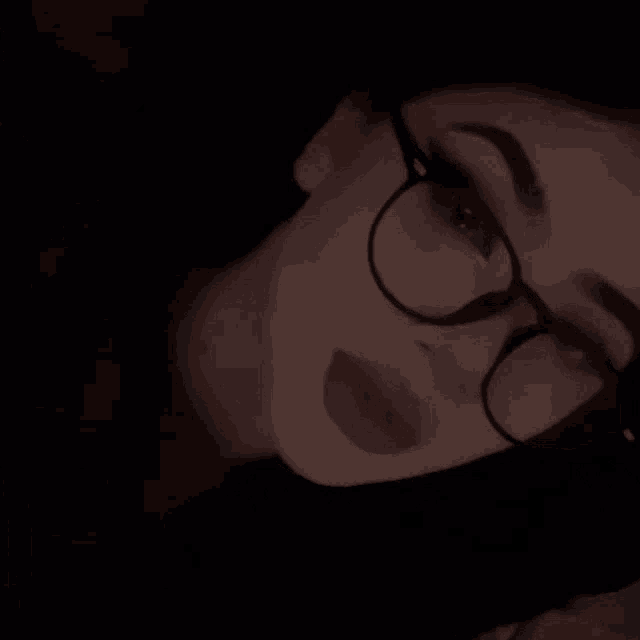 a woman wearing glasses and a black shirt is laying down in the dark .