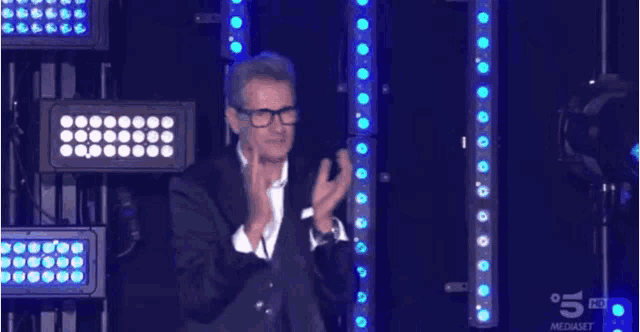 a man in a suit and glasses applauds while standing in front of a wall of lights
