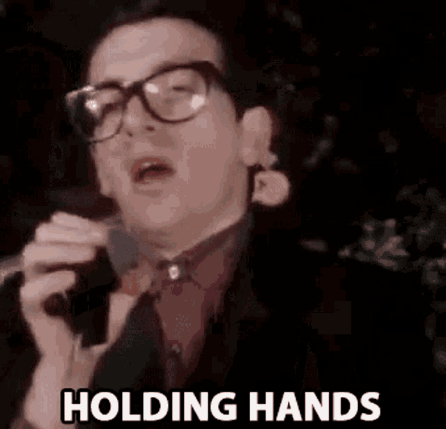 a man with glasses is singing into a microphone with the words holding hands below him .