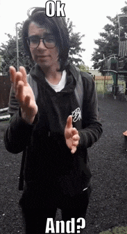 a man wearing glasses and a black hoodie is standing in a park with his hands in the air .