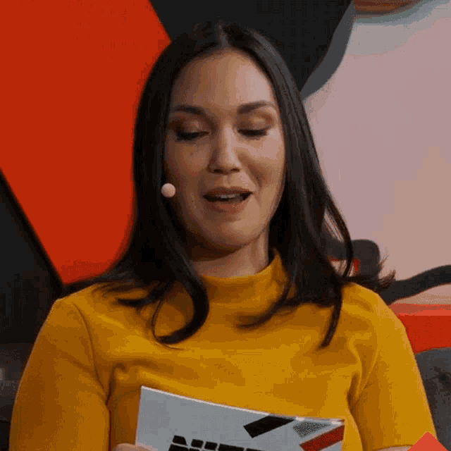 a woman wearing a yellow shirt and a microphone holds a piece of paper that says ' nbc ' on it