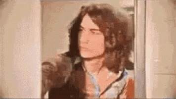 a man with long hair is standing in front of a door and looking at the camera .
