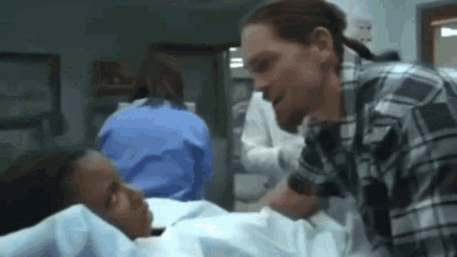 a man in a plaid shirt is talking to a woman laying in a hospital bed