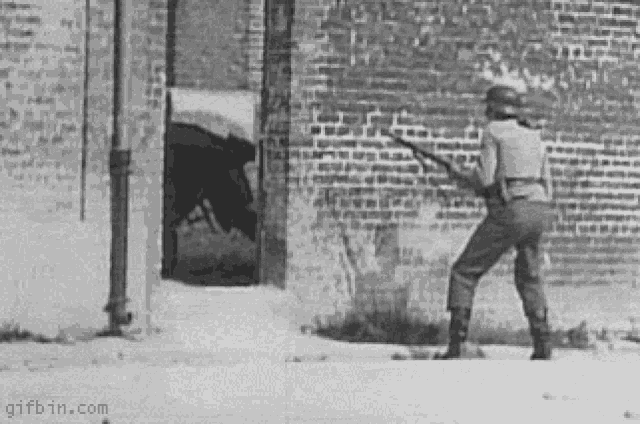 a black and white photo of a soldier holding a rifle in front of a brick wall .