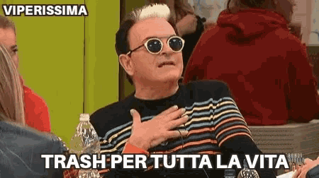 a man wearing sunglasses and a striped sweater is sitting at a table with his hands on his chest .