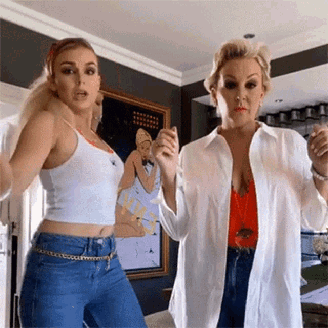 two women are dancing in a living room in front of a painting of a woman .