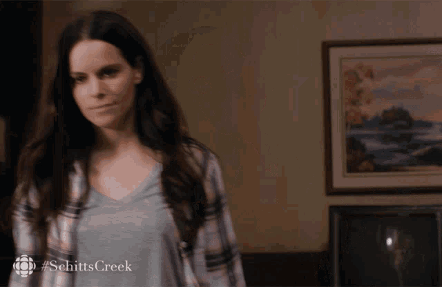 a woman in a plaid shirt is standing in front of a painting with #schitts creek written on the bottom