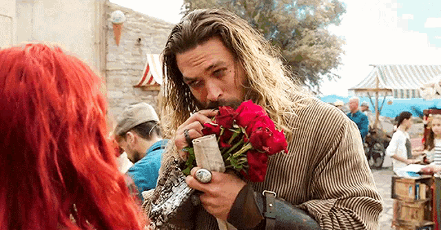 a man with long hair is holding a bouquet of red roses in his hand and smelling them .