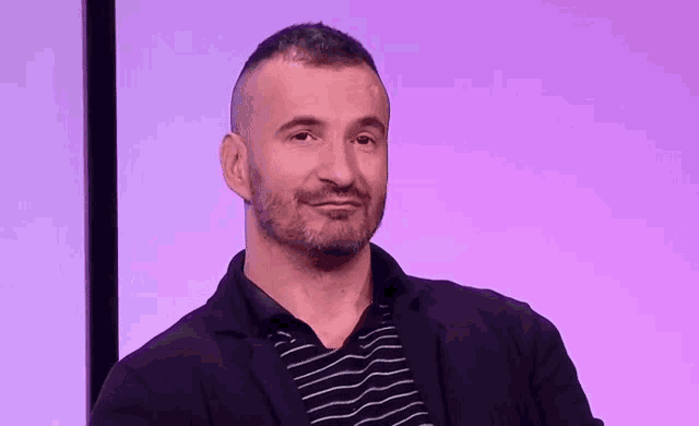 a man with a beard wearing a black jacket and striped shirt is smiling in front of a purple background .
