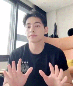 a young man wearing a black t-shirt is sitting in front of a window with his hands outstretched .