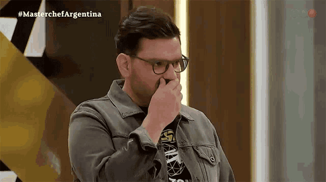 a man wearing glasses is covering his mouth with his hand while watching a show called master chef argentina