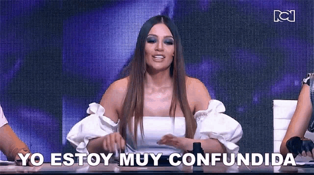 a woman in a white top is sitting at a table with the words yo estoy muy confundida written on the screen .