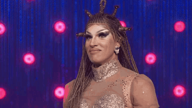 a drag queen with braids and a crown on her head stands on a stage