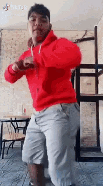 a man in a red hoodie and grey shorts is dancing in front of a table and chairs .
