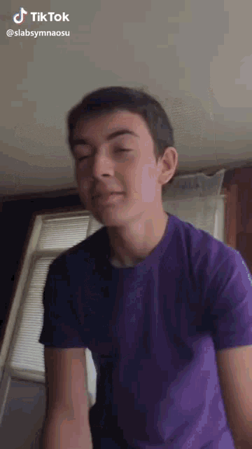 a young man in a purple shirt is standing in front of a window with his eyes closed .