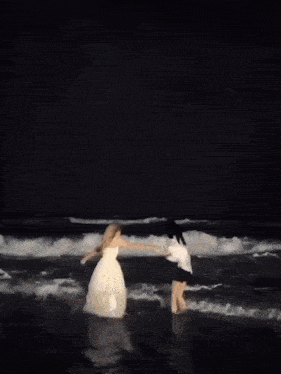 a woman in a white dress and a man in a white shirt are walking in the ocean at night