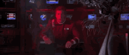 a man in a red uniform is standing in a room with a bunch of monitors .