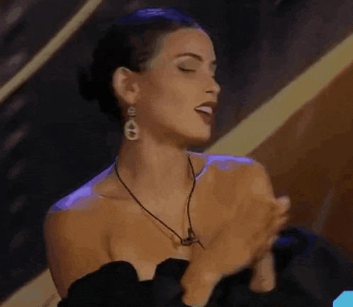 a woman in a black dress and earrings is making a heart shape with her hands .