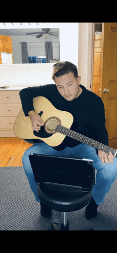 a man sits on a stool playing an acoustic guitar in front of a laptop