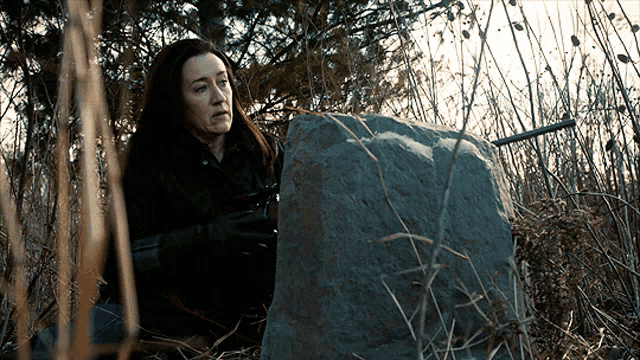 a woman is holding a gun while sitting next to a large rock