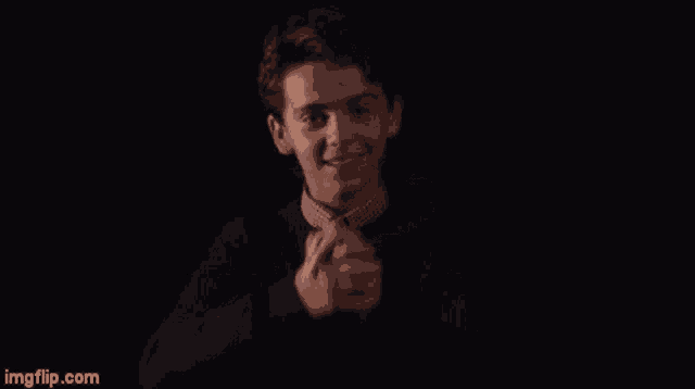a young man in a suit and tie is smiling in the dark while holding his hands together .