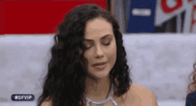 a woman with curly hair is wearing a necklace and looking at the camera with her eyes closed .