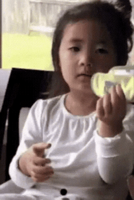 a little girl in a white shirt is holding a bottle and making a face