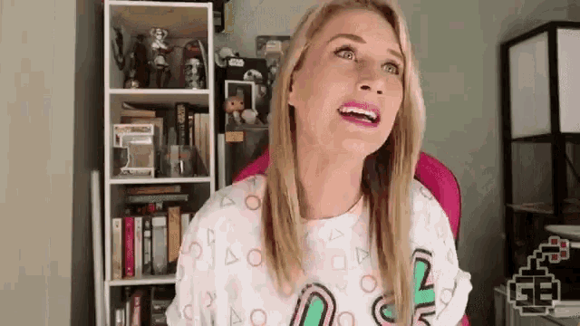 a woman is sitting in a pink chair in front of a bookshelf and making a funny face .