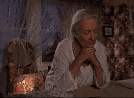 an elderly woman is sitting at a table with her hands folded in prayer
