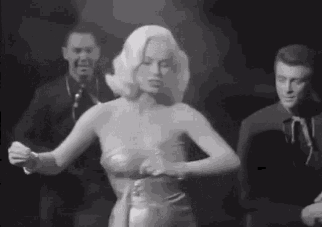 a black and white photo of a woman in a dress dancing with two men in the background .
