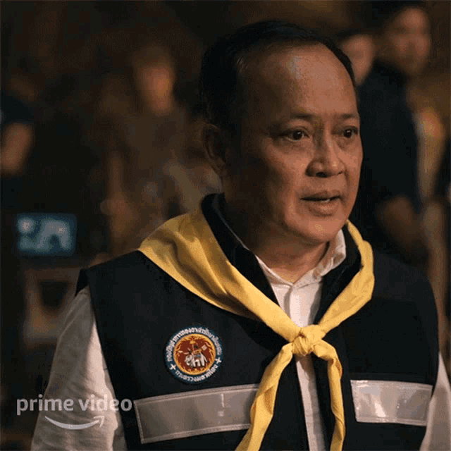 a man with a yellow scarf around his neck has a patch on his vest that says " prime video "