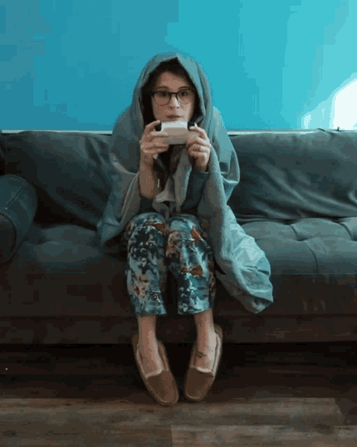 a woman wrapped in a blanket sits on a couch holding a video game controller