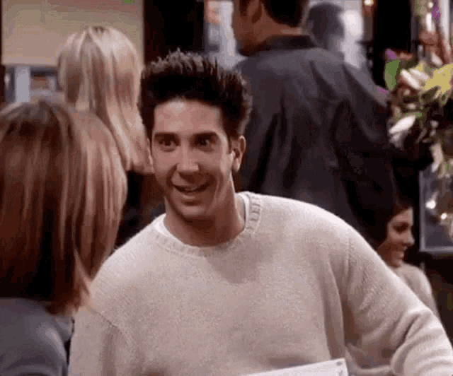 a man in a sweater is smiling while standing in front of a crowd of people .