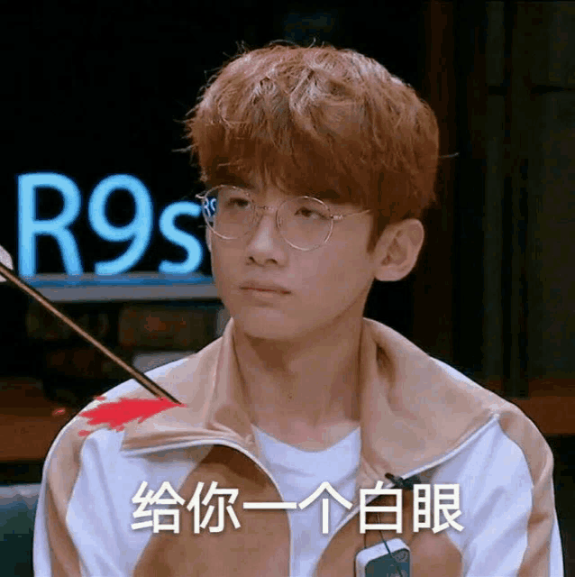 a young man wearing glasses is sitting in front of a sign that says r9s .