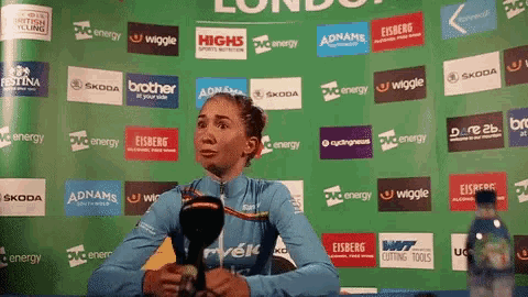 a woman speaking into a microphone in front of a wall of advertisements including adnams