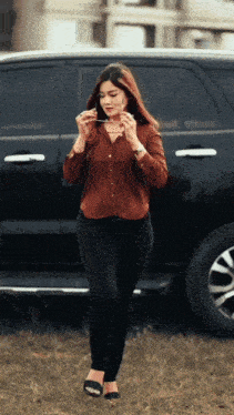 a woman in a brown shirt and black jeans is standing in front of a black suv
