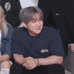 a man in a blue t-shirt is sitting on a couch with his hands folded in front of a group of people .