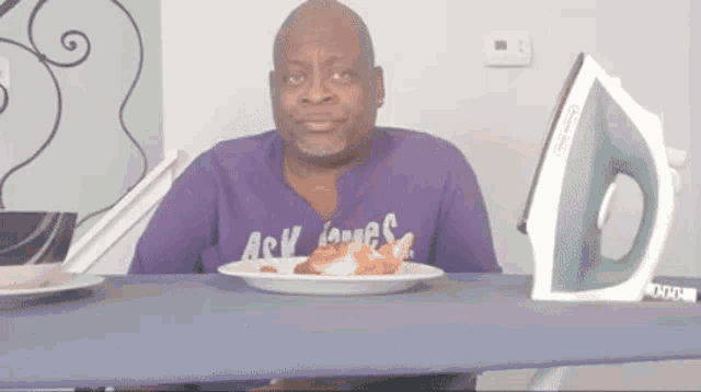 a man in a purple shirt is sitting at a table with a plate of food and an iron ..