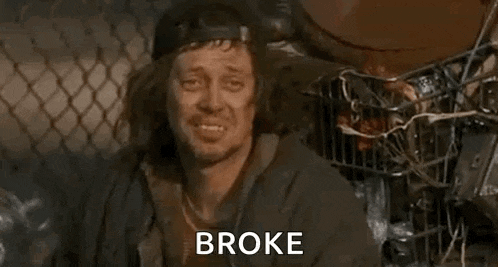 a homeless man is sitting in front of a chain link fence with the words `` broke '' written on his face .