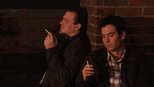two men are leaning against a brick wall one is looking at the camera
