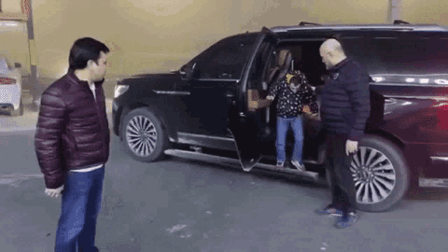 a man in a red jacket stands next to a man in a black jacket standing next to a black suv